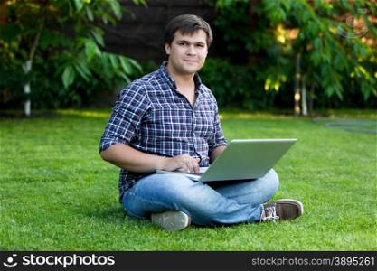 Young smiling man sitting on grass and working with laptop