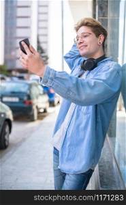 Young smiling man takes a photo of himself while resting on a glass window on an out of focus background. Lifestyle concept.. Young man takes a selfie while posing