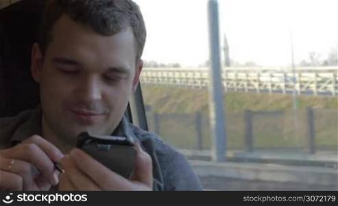 Young smiling man traveling by train and using smart phone. He sending message and looking out the window