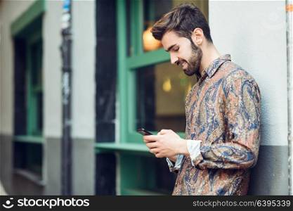 Young smiling man wearing casual clothes looking at his smartphone in the street. Guy with beard and modern hairstyle in urban background
