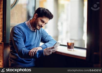 Young smiling man with blue sweater looking at his smartphone in a modern pub. Bearded guy with modern hairstyle drinking a cola.