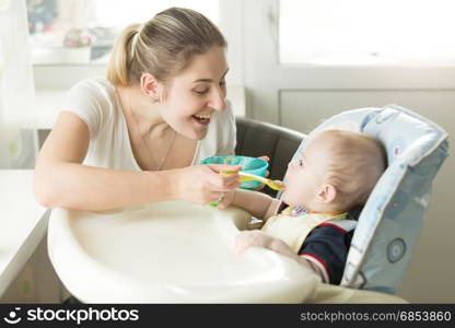 Young smiling mother giving apple sauce to her baby in highchair