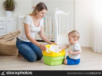 Young smiling mother sitting on floor at living room and teaching her baby boy how to use chamber pot
