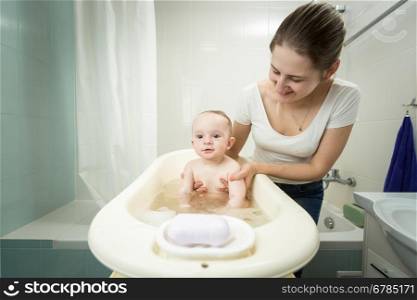 Young smiling mother washing her cute baby in plastic bath at bathroom