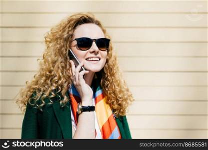 Young smiling office worker in sunglasses having blonde curly hair talking on cell phone during a break standing at street against beige background with copy space for your promotional content