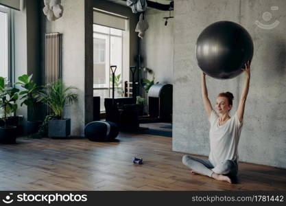 Young smiling red haired woman exercising in fitness studio, holding large silver exercise ball with both hands above her head and enjoying healthy lifestyle while sitting in lotus position on floor. Smiling woman in lotus position enjoying pilates or fitness workout indoors