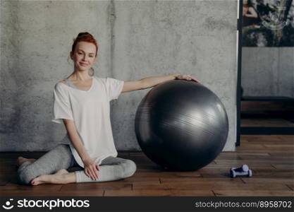 Young smiling red haired woman posing with large exercise pilates fitball while sitting on floor against grey wall in fitness studio, relaxing after training. Healthy and active lifestyle concept. Happy young fitness woman relaxing after pilates workout while sitting with large exercise ball on floor