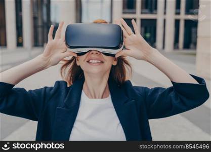 Young smiling redhead female entrepreneur testing portable VR goggles, businesswoman interacting with virtual reality and expressing positive emotions while standing alone on city street. Young smiling redhead female entrepreneur testing portable VR goggles on city street
