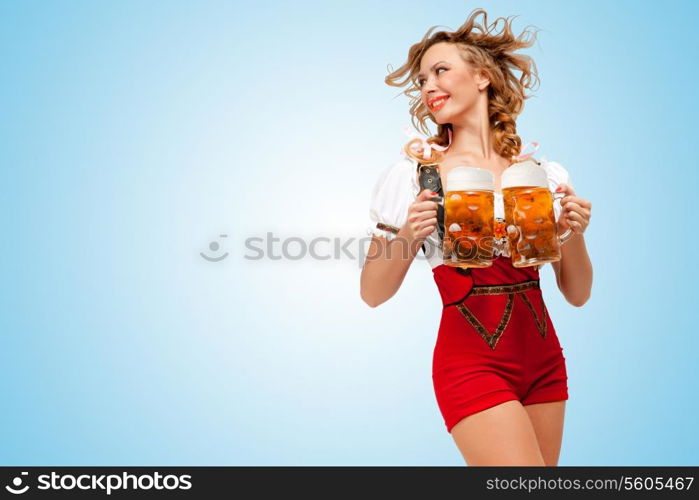 Young smiling sexy Swiss woman wearing red jumper shorts with suspenders in a form of a traditional dirndl, holding two beer mugs and looking aside on blue background.