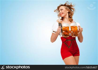 Young smiling sexy Swiss woman wearing red jumper shorts with suspenders in a form of a traditional dirndl, holding two beer mugs and looking aside on blue background.