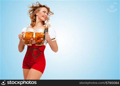 Young smiling sexy Swiss woman wearing red jumper shorts with suspenders in a form of a traditional dirndl, holding two beer mugs and looking aside on blue background.