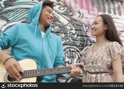 Young smiling street musician leaning on a wall with graffiti drawings, playing his guitar, and flirting with a young woman in a dress