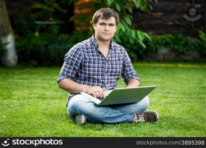 Young smiling student sitting on grass at park and typing on laptop