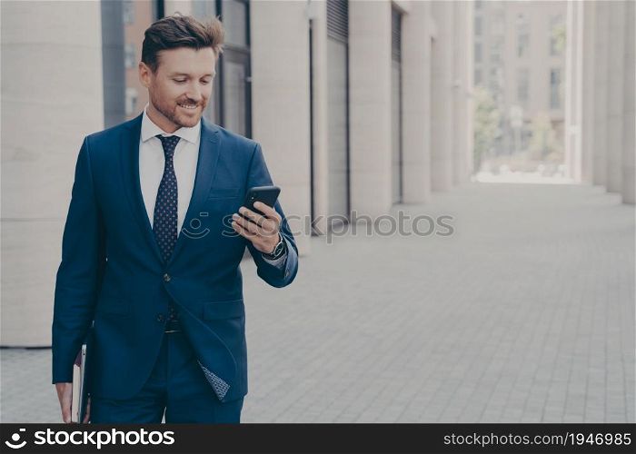 Young smiling successful male ceo executive looking at smartphone with satisfied face expression, reading news, browsing internet while going at business meeting with client, standing outdoors. Young smiling successful male ceo executive looking at smartphone with satisfied expression outside