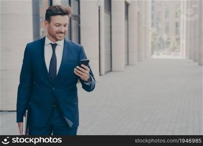 Young smiling successful male ceo executive looking at smartphone with satisfied face expression, reading news, browsing internet while going at business meeting with client, standing outdoors. Young smiling successful male ceo executive looking at smartphone with satisfied expression outside