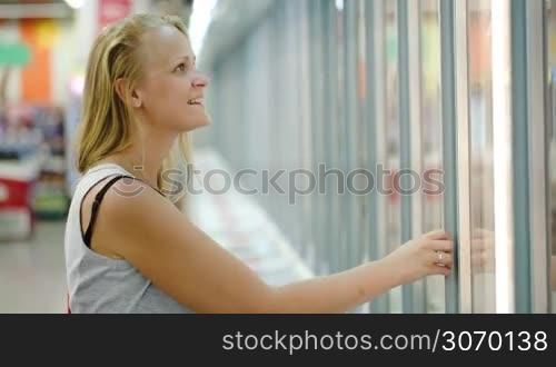 Young smiling woman coming up to the fridge in the shop and taking product from it