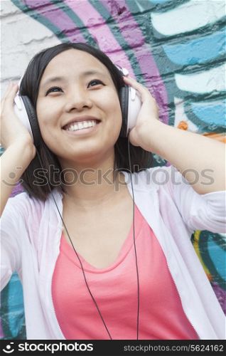Young smiling woman holding her headphones while enjoying listening to music in front of wall with graffiti