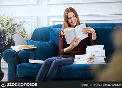 Young smiling woman is reading on a sofa with a pile of books.