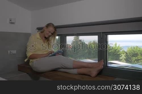 Young smiling woman is sitting on window sill and typing in her tablet.