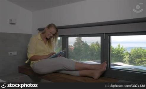 Young smiling woman is sitting on window sill and typing in her tablet.