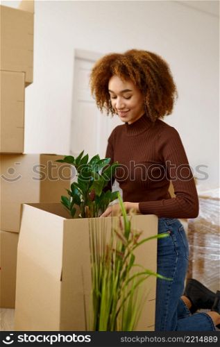 Young smiling woman packing flower into cardboard box. Moving day after buying real estate. Young woman packing flower during moving day