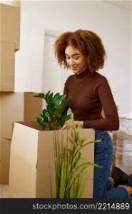Young smiling woman packing flower into cardboard box. Moving day after buying real estate. Young woman packing flower during moving day
