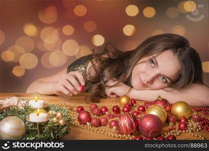Young smiling woman sits, relaxed, on a wooden table, surrounded by Christmas decorations and candles, thinking and playing with globes.