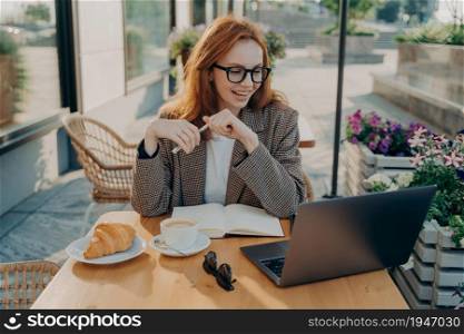 Young smiling woman sitting at cafe table outdoors and watching online lesson or webinar on laptop, happy redhead female in eyeglasses studying remotely, making some notes during distant education. Young smiling woman sitting at cafe table outdoors and watching online lesson or webinar on laptop
