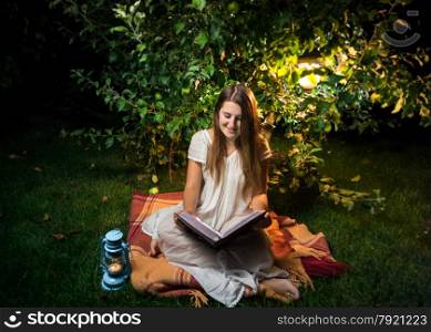 Young smiling woman sitting at night in garden and reading big old book