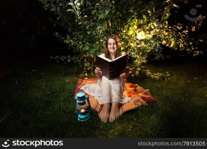 Young smiling woman sitting on grass at night and reading big old book