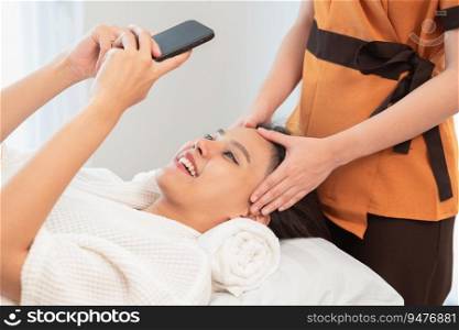 Young smiling woman taking selfie at beauty salon. Beautiful happy girl using smartphone while having spa facial massage.