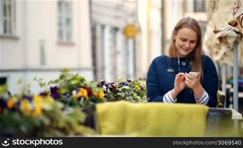 Young smiling woman typing text message on her cell phone sitting alone in outdoor cafe
