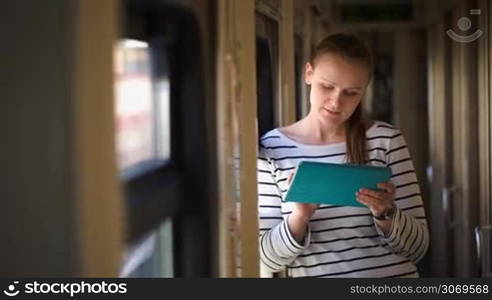 Young smiling woman using tablet PC standing near the window in train hallway