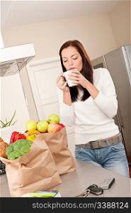 Young smiling woman with groceries drinking cup of coffee in the kitchen