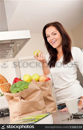 Young smiling woman with groceries holding apple in the kitchen