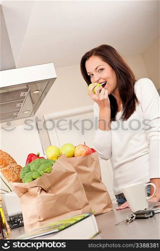 Young smiling woman with groceries in the kitchen biting apple