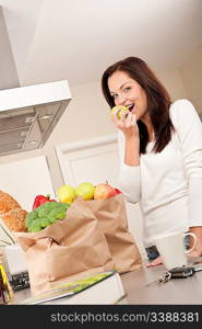 Young smiling woman with groceries in the kitchen biting apple