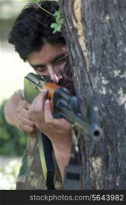Young soldier aiming with rifle while standing behind tree