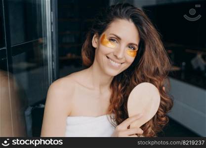 Young spanish girl with under eye patches brushes long hair with wooden hairbrush after shower, smiling pretty female doing morning beauty treatment routine, combing hairs, moisturizing skin at home.. Young spanish girl with under eye patches brushes hair after shower. Morning beauty routine at home