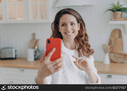 Young spanish woman has kitchen blog. Lady with mobile telephone in her stylish kitchen at home looking at camera and making video. Modern white scandinavian interior. Stove, worktop and cuisine.. Young woman has kitchen blog. Lady with telephone at home looking at camera and making video.