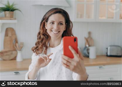 Young spanish woman has kitchen blog. Lady with mobile telephone in her stylish kitchen at home looking at camera and making video. Modern white scandinavian interior. Stove, worktop and cuisine.. Young woman has kitchen blog. Lady with telephone at home looking at camera and making video.