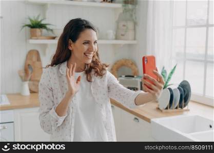 Young spanish woman has video call on phone at kitchen. Girl is looking at camera, waving hand and smiling. White scandinavian interior. Daylight through the window.. Young spanish woman has video call on phone at kitchen. Girl is waving hand and smiling.