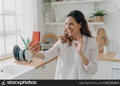 Young spanish woman has video call on phone at kitchen. Girl is looking at camera, waving hand and smiling. White scandinavian interior. Daylight through the window.. Young spanish woman has video call on phone at kitchen. Girl is waving hand and smiling.