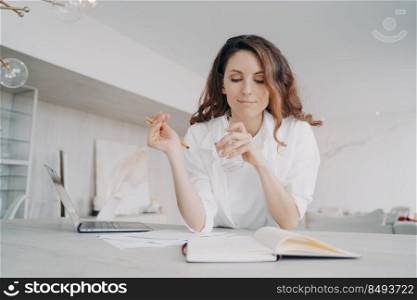 Young spanish woman in white is taking notes. Teacher is checking schedule. Girl is writing down into notebook at the desk preparing to online lesson. Remote work and study from home on quarantine.. Young spanish woman teacher in white is taking notes. Remote work and study at home.