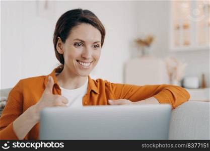 Young spanish woman is speaking online and showing thumb. Girl has online conference indoors. Lady with laptop is talking at internet meeting. Working on a couch at home while quarantine.. Young spanish woman is speaking online and showing thumb. Girl has online conference at home.