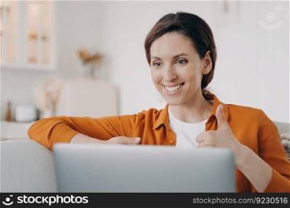 Young spanish woman is speaking online and showing thumb. Girl has online conference indoors. Lady with laptop is talking at internet meeting. Working on a couch at home while quarantine.. Young spanish woman is speaking online and showing thumb. Girl has online conference at home.