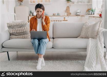 Young spanish woman is talking on phone and holding laptop. Girl has consultation on phone. Businesswoman is sitting on sofa at home holding computer. Remote work on quarantine.. Young spanish woman is talking on phone and holding laptop. Remote work on quarantine.