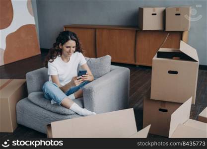 Young spanish woman posting news. Happy apartment buyer unpacking boxes. Girl is moving to new home. Relaxed young lady clicks mobile phone. Independence and achievement concept.. Young spanish woman posting news. Happy apartment buyer unpacking boxes.