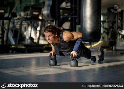 Young sportsman doing push-up exercise with kettlebell during training at gym. Workout and bodybuilding concept. Young sportsman doing push-up exercise with kettlebell during training at gym