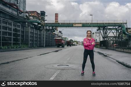 Young sportswoman posing arms crossed outdoors on a rainy day in an industrial zone. Sportswoman posing arms crossed in an industrial zone